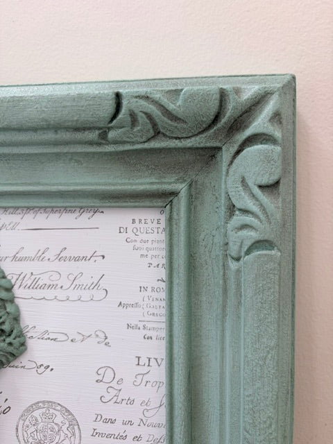A close-up of a distressed sage green ornate picture frame with carved scrollwork corners, mounted on a light pink wall.