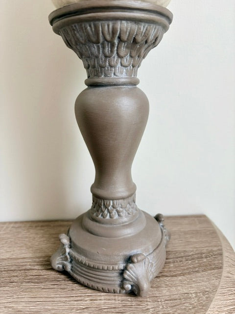 A distressed brown candlestick holder with ornate classical detailing, shown resting on a wooden surface against a plain off-white wall.