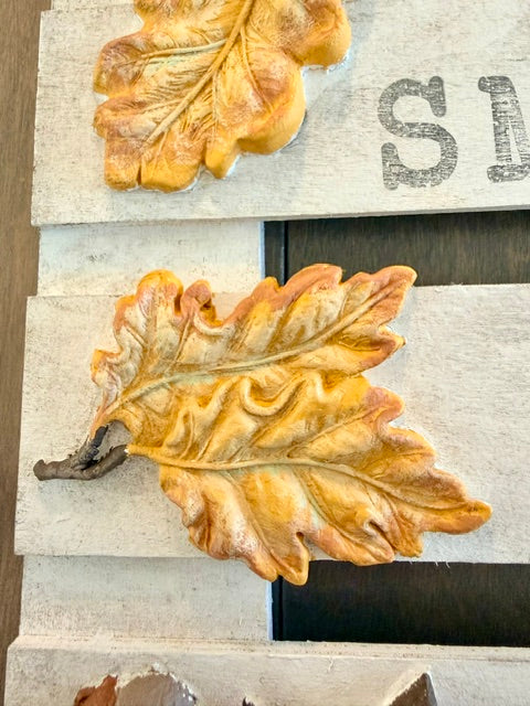 Two decorative 3D oak leaves in autumn shades of orange, yellow, and brown, mounted on a rustic white wooden plank with a dark wood slat in the background.