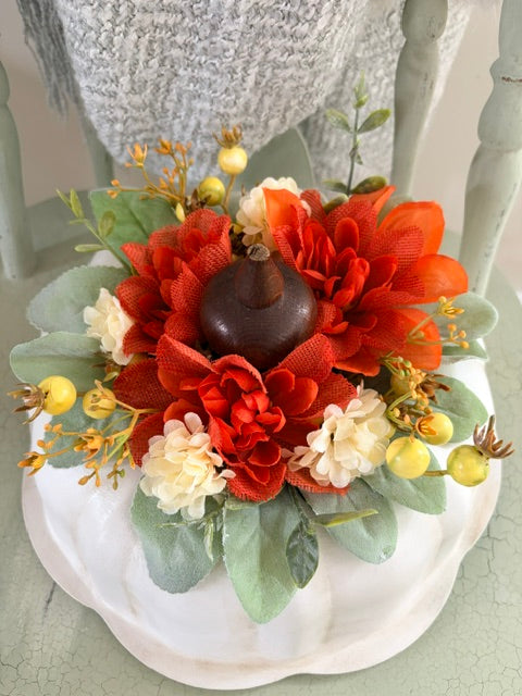An autumn-themed floral arrangement featuring vibrant orange and red artificial flowers, sage green leaves, small yellow berries, and a dark brown gourd centerpiece, displayed on a white scalloped plate.