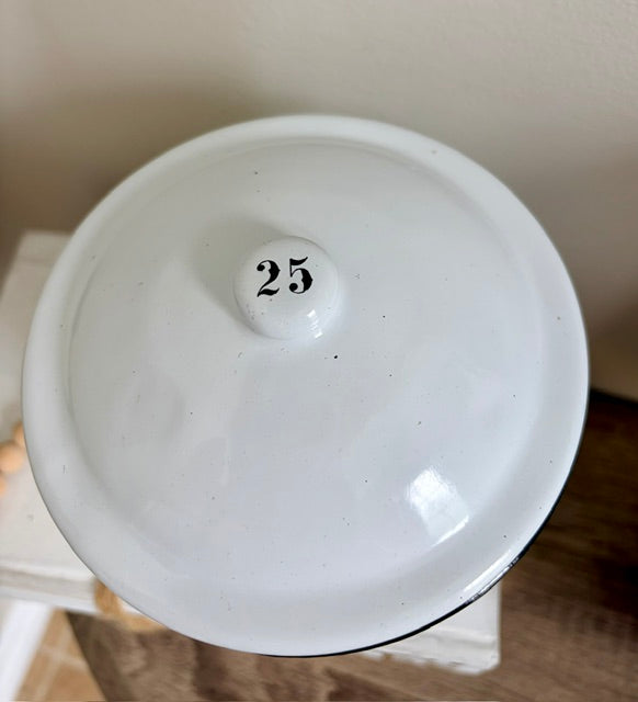A close-up of a white ceramic lidded container or jar with a small knob in the center printed with the number 25, resting on a wooden surface against a neutral wall.