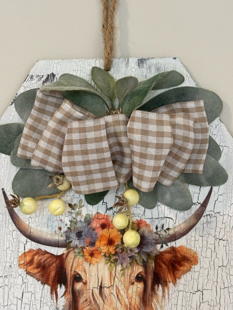 Decorative wall hanging with a checkered bow, flowers, and cow skull design on a textured background.
