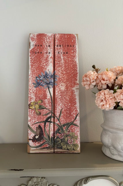 Decorative wooden panel with floral designs and text on a shelf with flowers in the background.