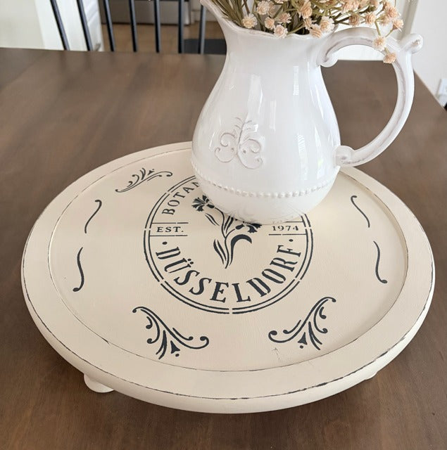Round wooden tray with decorative text and a white pitcher on a wooden surface