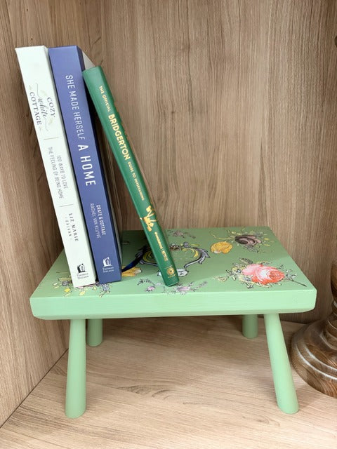 Small green stool with floral design holding books against a wooden wall.