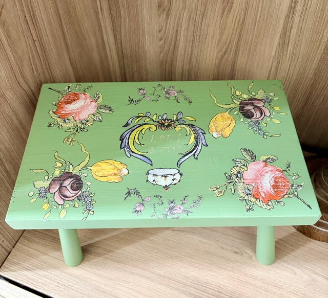 Green step stool with floral and decorative designs on a wooden floor.