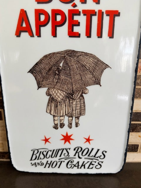 Decorative cutting board with a design of two people under an umbrella, text 'Bon Appétit', and food items below on a white background.