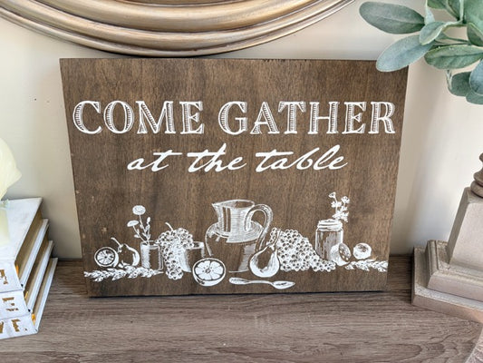 Wooden sign with 'Come Gather at the Table' text and illustrations on a wooden surface.