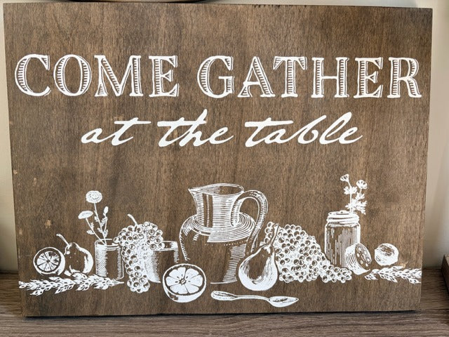 Wooden sign with white text 'Come Gather at the Table' and illustrations of a pitcher, jar, and fruits.