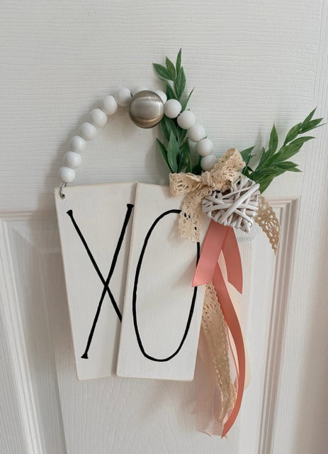 Decorative wall hanging with 'XO' sign, beads, and ribbons on a white background