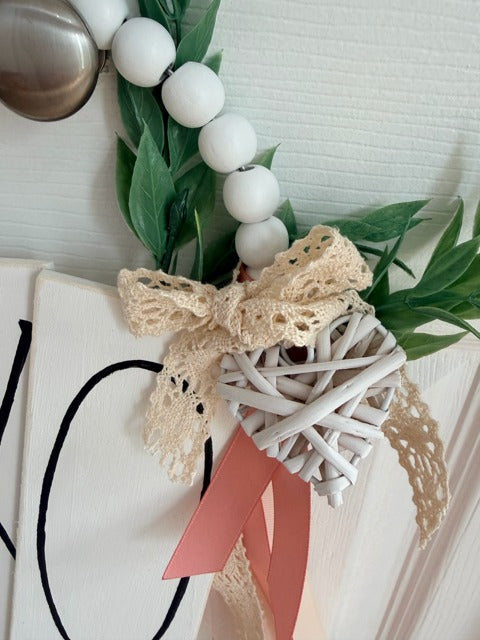 Decorative heart with white balls, green leaves, and a pink ribbon on a white surface.