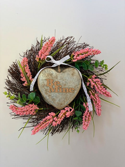 Decorative wreath with pink flowers and a heart-shaped metal with 'Be Mine' text on a light background