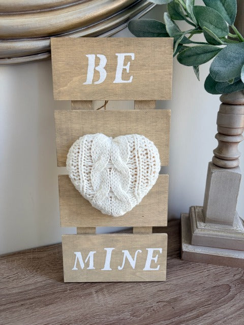Wooden sign with knitted heart and 'BE MINE' text on a wooden surface.
