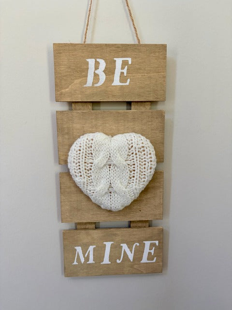 Wooden wall hanging with knitted heart and text 'BE MINE' on a plain background