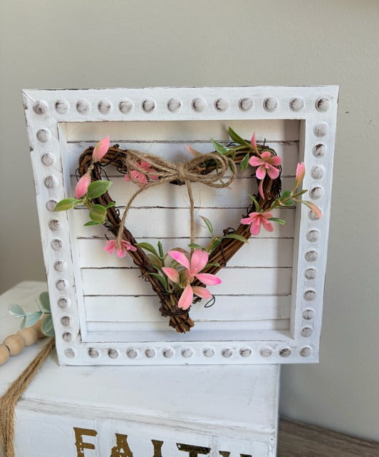Decorative heart made of grapevine and pink flowers in a white wooden frame.