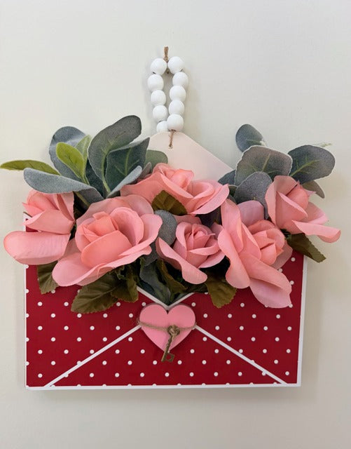 Decorative wall hanging with pink flowers, green leaves, and a red envelope with white polka dots on a light gray background.