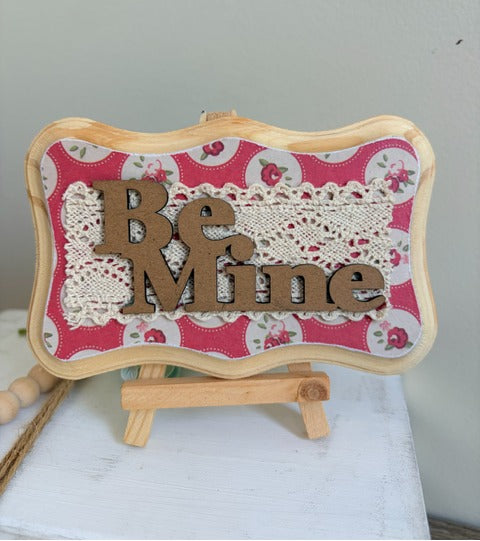 Decorative sign with 'Be Mine' on a floral background with a wooden stand.