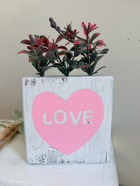 White wooden block with a pink heart and 'LOVE' text, featuring red plants 
