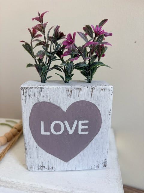 Decorative wooden block with a heart and 'LOVE' text, holding artificial flowers.