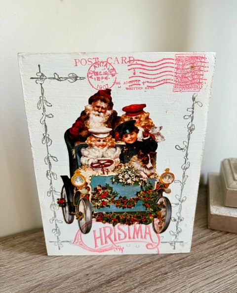 Vintage-style Christmas wood postcard with Santa Claus and chidren in a car, featuring decorative elements and a red postmark.