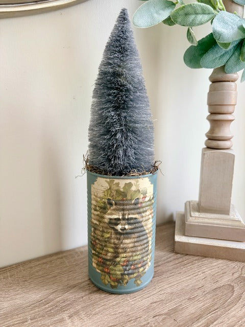 Decorative bottle brush tree in a cylindrical container with a raccoon design