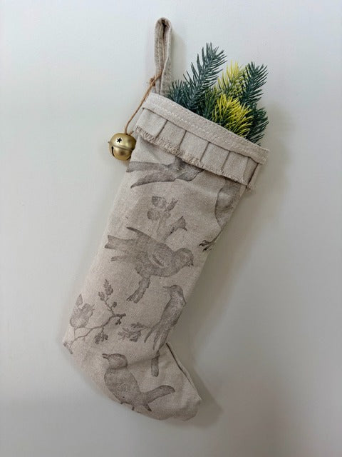 Beige Christmas stocking with bird design, gold bell, and greenery on a white background