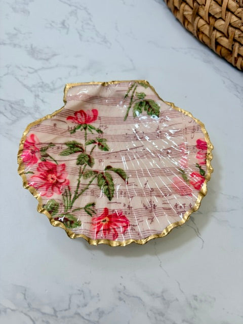 A small decorative dish with an irregular scalloped edge, featuring a floral  pattern on a pink music note background with a gold-toned distressed rim.