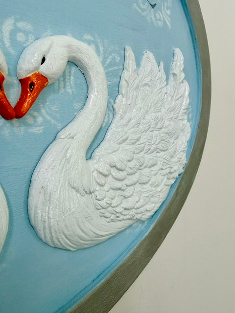 A close-up of a decorative wall plaque featuring a raised white swan with an orange beak on a light blue background, with part of another swan visible at the left edge.
