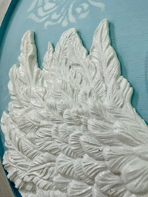 A close-up of a white textured wing or feather-shaped decorative element with raised leaf-like details, set against a light blue background.