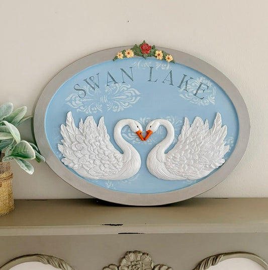 An oval decorative wall plaque featuring two white swans facing each other with their necks forming a heart shape, on a light blue background with the words 'SWAN LAKE' and small flowers at the top. The plaque has a gray border and is displayed on a shelf against a light wall.