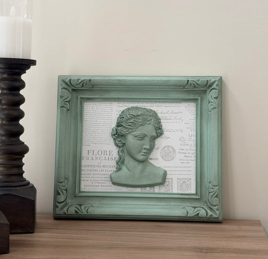 A framed decorative wall art piece with a pale green ornate frame, featuring a sculpted classical bust over a vintage-style printed background, displayed on a wooden surface against a light wall.