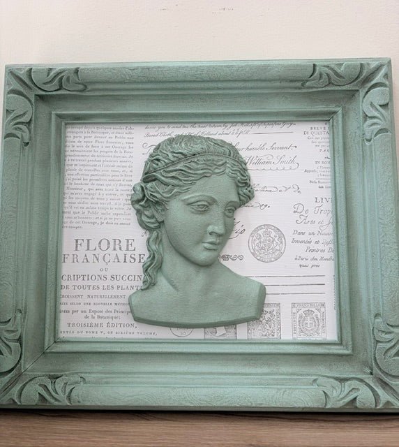 A decorative framed wall art piece featuring a pale green classical bust sculpture mounted over a vintage-style French script and postage stamp background, all enclosed in an ornate mint green frame.