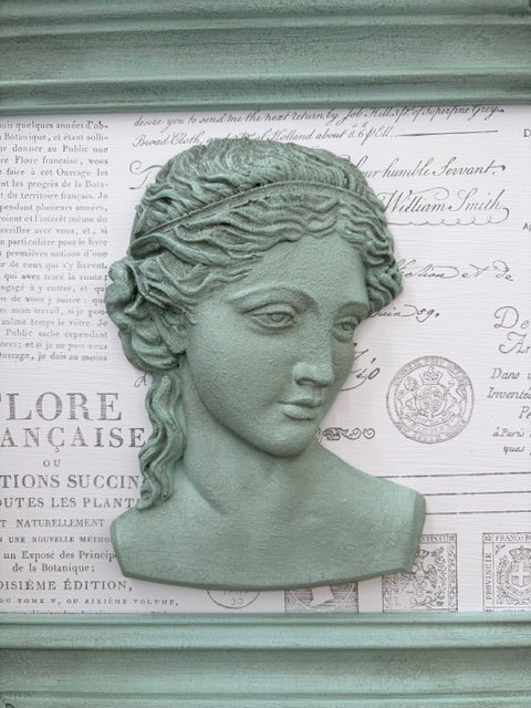 A green-gray resin classical bust sculpture of a woman with braided hair, mounted on a vintage-style printed paper background.