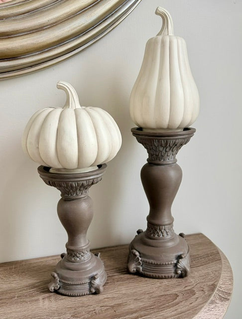 Two decorative faux pumpkins in an off-white finish, displayed on ornate dark brown candlesticks on a wooden surface against a neutral wall.