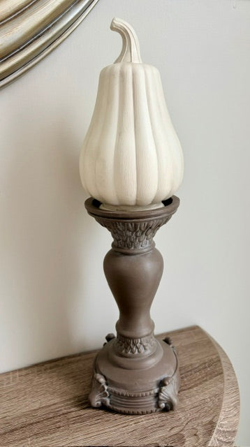 A decorative faux pumpkin in an off-white color sits on an ornate, dark brown pedestal candle holder, placed on a wooden surface against a neutral wall.