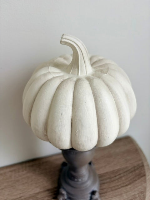 A white faux pumpkin decorative ornament with a ribbed texture and curved stem, displayed on a dark gray pedestal stand over a wooden surface against a plain light gray wall.