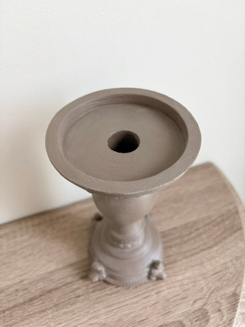 A single matte brown candlestick holder with a circular top and pedestal base, photographed on a light wood surface against a plain off-white wall.
