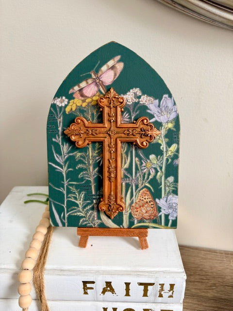 A decorative arched plaque with a green background, floral and butterfly illustrations, and a large ornate resin cross mounted in the center, displayed on a small stand atop stacked white books.