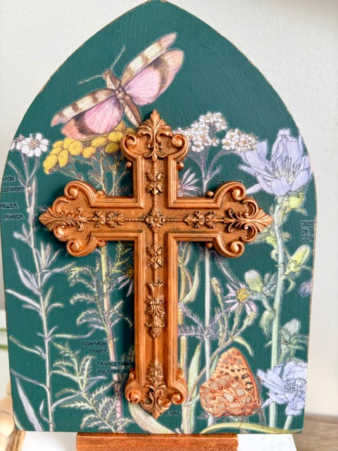 An ornate carved resin cross in a warm copper-brown tone, mounted on a decorative arched panel featuring botanical illustrations, flowers, and butterflies on a dark green background.