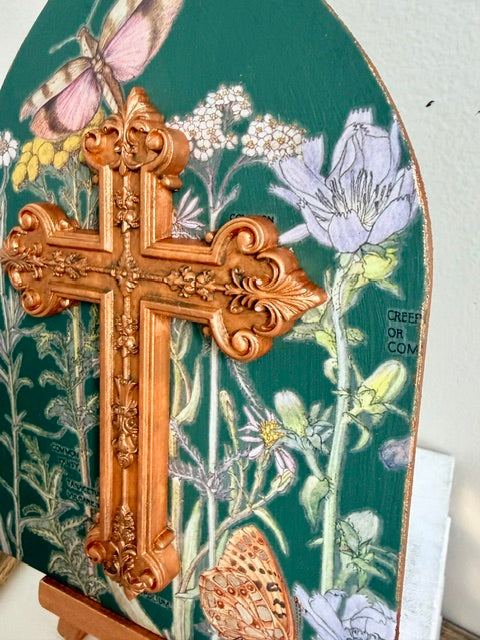 An ornate carved resin cross in a warm coppery gold finish, mounted on a decorative green panel featuring painted flowers, butterflies, and delicate botanical illustrations.