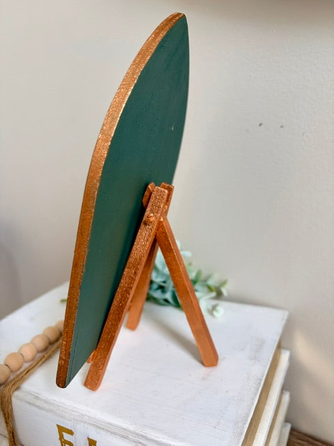 A small decorative tabletop sign with a dark green oval board and a natural wood A-frame stand, displayed on stacked white books against a light wall.
