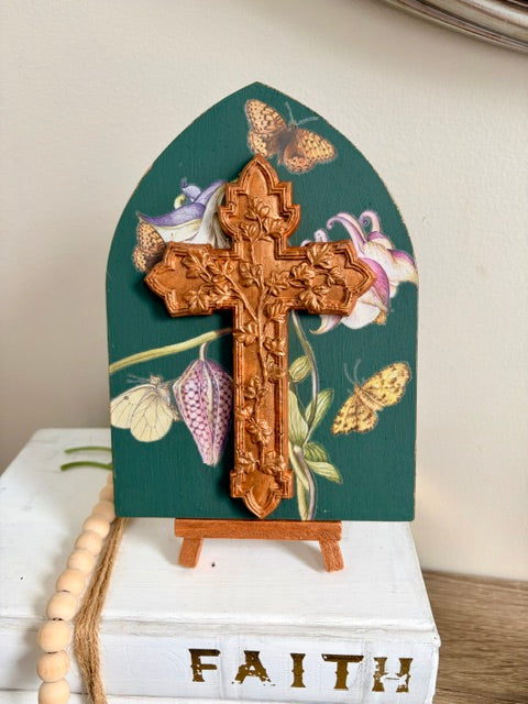A decorative resin cross with an ornate carved design, mounted on a dark teal painted arched plaque featuring butterflies and flowers. The plaque is displayed on a small wooden easel stand resting on a white book with the word 'FAITH' on the front.