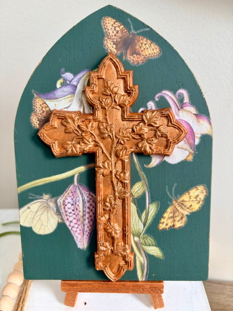 An arched decorative plaque with a carved resin cross mounted on a dark teal background featuring painted flowers and butterflies, displayed on a small stand.