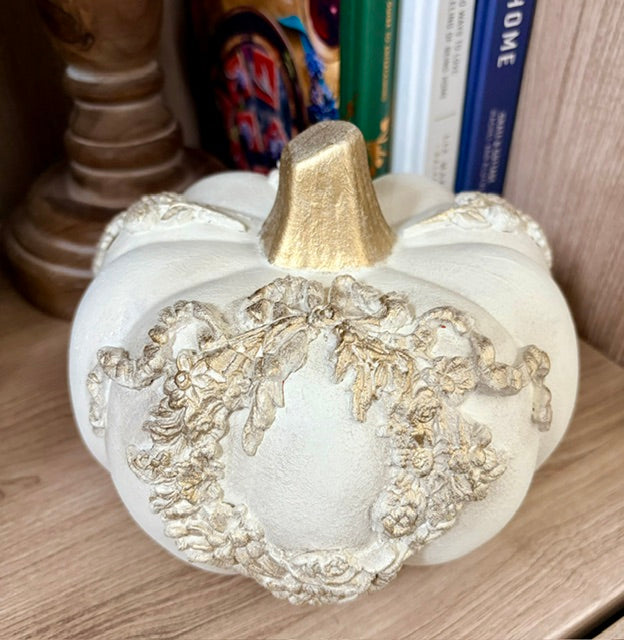 A decorative faux pumpkin in off-white with a metallic gold top and a raised gold floral wreath design on the front, displayed on a wooden shelf.