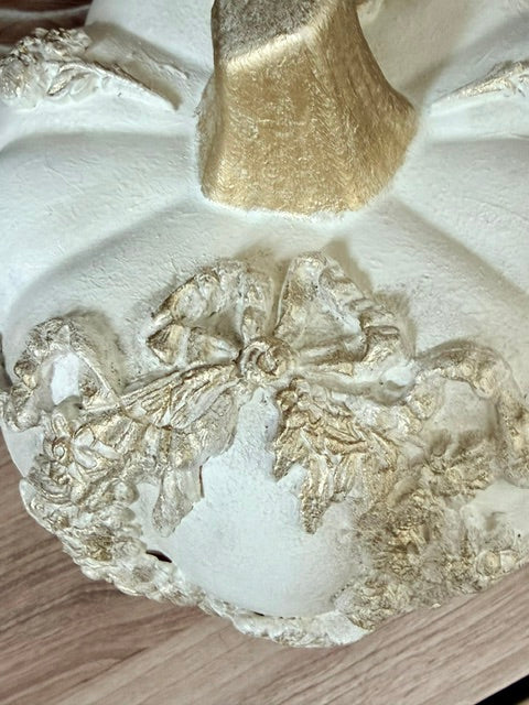 A close-up of a decorative pumpkin with a textured off-white finish and metallic gold leaf detailing on the stem and carved floral motifs.