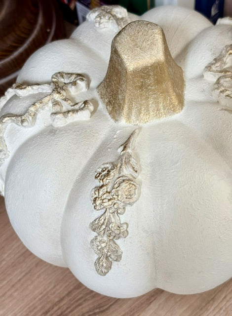 A white decorative pumpkin with a textured finish, featuring a metallic gold top stem and gold floral vine accents, resting on a light wood surface.