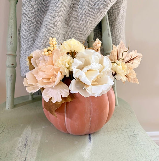 A decorative faux floral arrangement in a ribbed peach pumpkin vase, placed on a distressed sage green chair with a gray knit throw draped behind it.