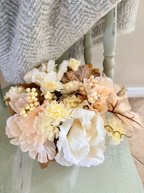 An artificial floral arrangement featuring cream, white, and peach peonies, dried leaves, and pearl-like berries, styled on a light green table with a gray knit blanket draped over a chair.