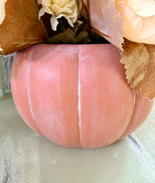 A terracotta faux pumpkin vase filled with artificial autumn leaves and cream-colored flowers, set on a light-colored surface.
