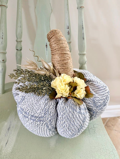 A decorative knitted pumpkin in a heathered light blue and white pattern, styled with a natural jute-wrapped stem, cream-colored faux flowers, and dried botanicals, displayed on a distressed mint green chair.
